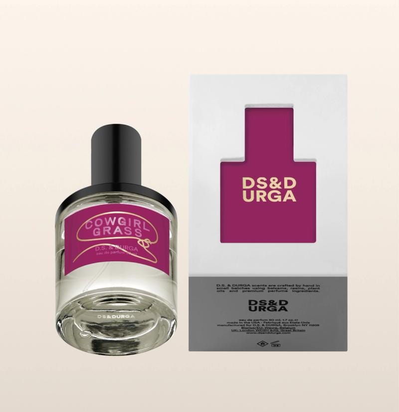 A clear glass perfume bottle labeled "Cowgirl Grass" by DS & DURGA sits beside its white and purple box packaging. The bottle has a black cap and the packaging features a cutout design revealing the bottle's label.