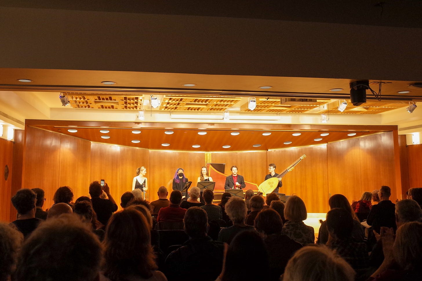 An audience faces a stage paneled in wood. Five performers are visible with a harpsichord in the background, and a theorbo is held by the right-most person on stage.