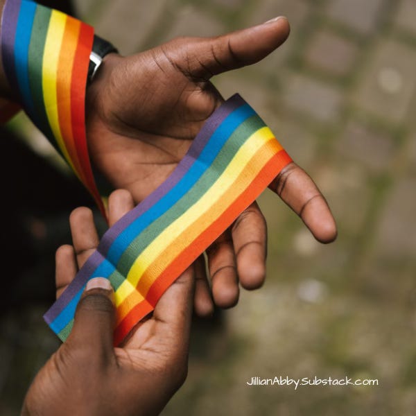 Two Black hands, wearing a suit and watch, and holding a Pride colored ribbon.