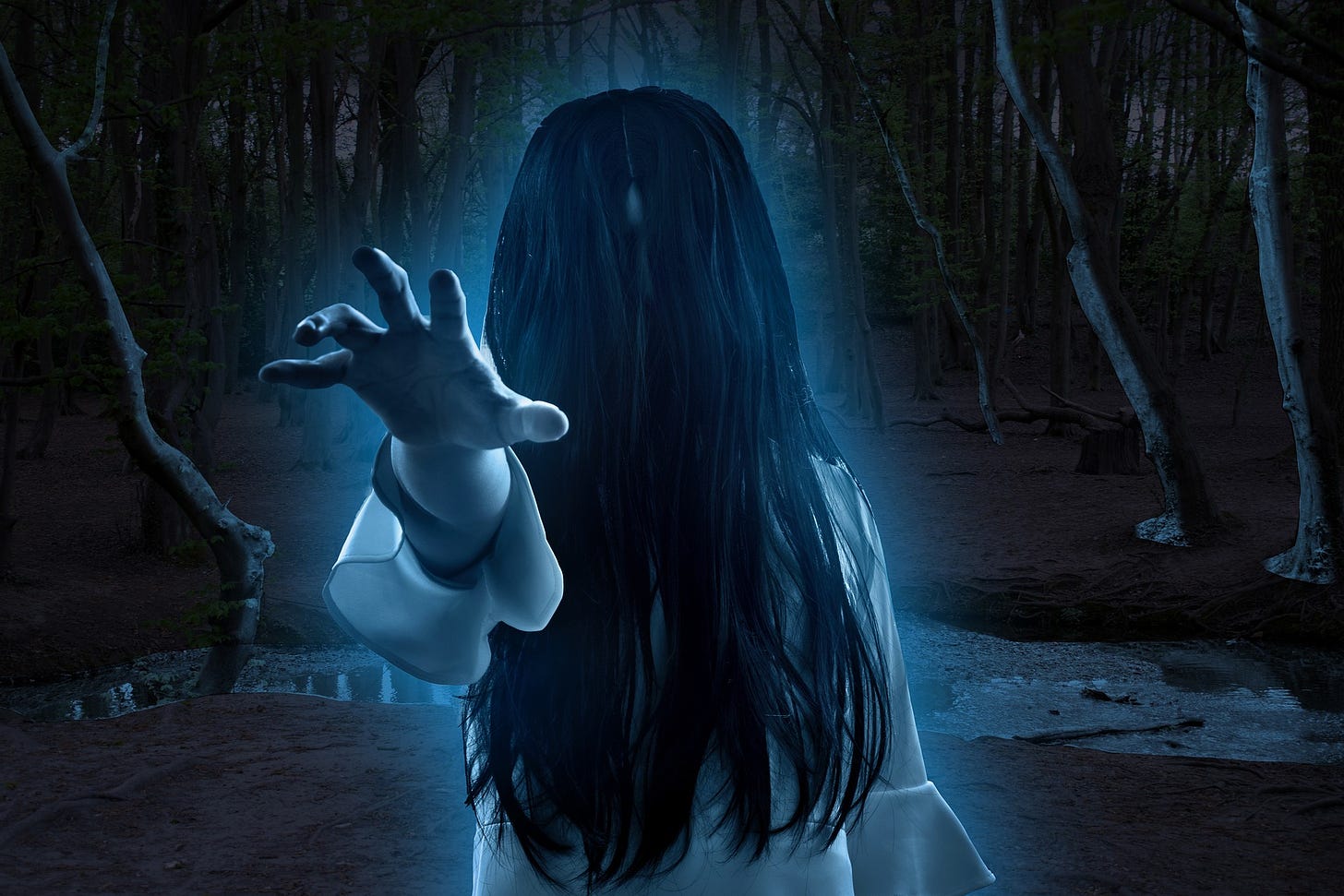 Scary woman in the woods at night. Her long hair covers her face and her arm is reached out as if to grab what's in front of her.