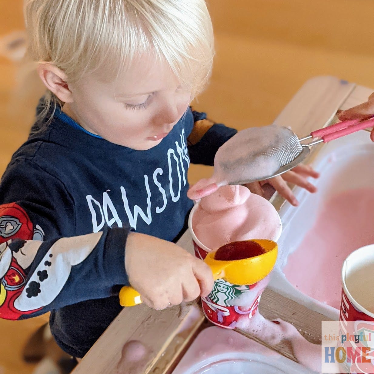 a blonde toddler scoops pink foam into a large red Starbucks cup