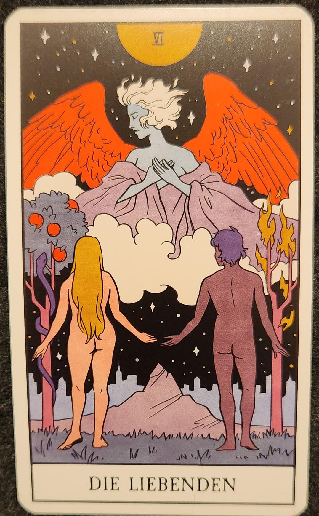 A tarot card labelled V Die Libenden. Two naked people, a man and a woman, stand facing an angel hanging in a starry night sky. Their hands are held out towards each other, but not touching. The angel is a blue person with short, white hair and bright red wings. Their eyes are closed and their hands are clasped over their heart. To the left of the woman is an apple tree, covered in red apples. To the left of the man is a bare tree whose branches are on fire. 