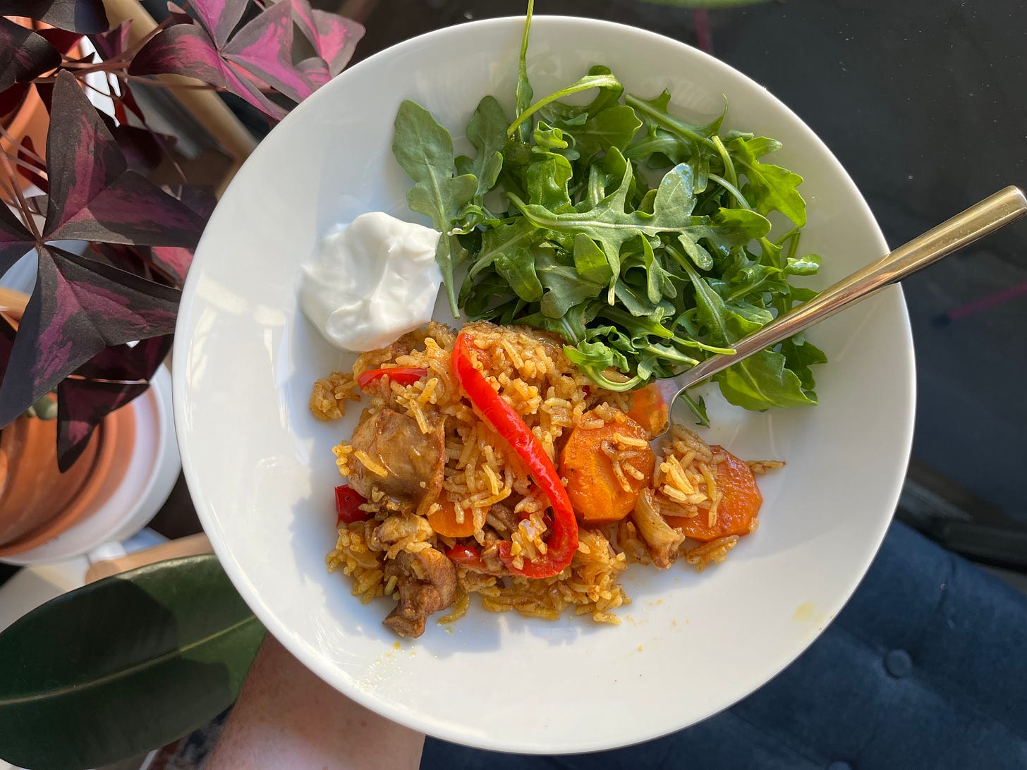 A plat with kabsa chicken and rice with red bell pepper and slices of carrot visible, a side of arugula, and a dollop of yogurt.