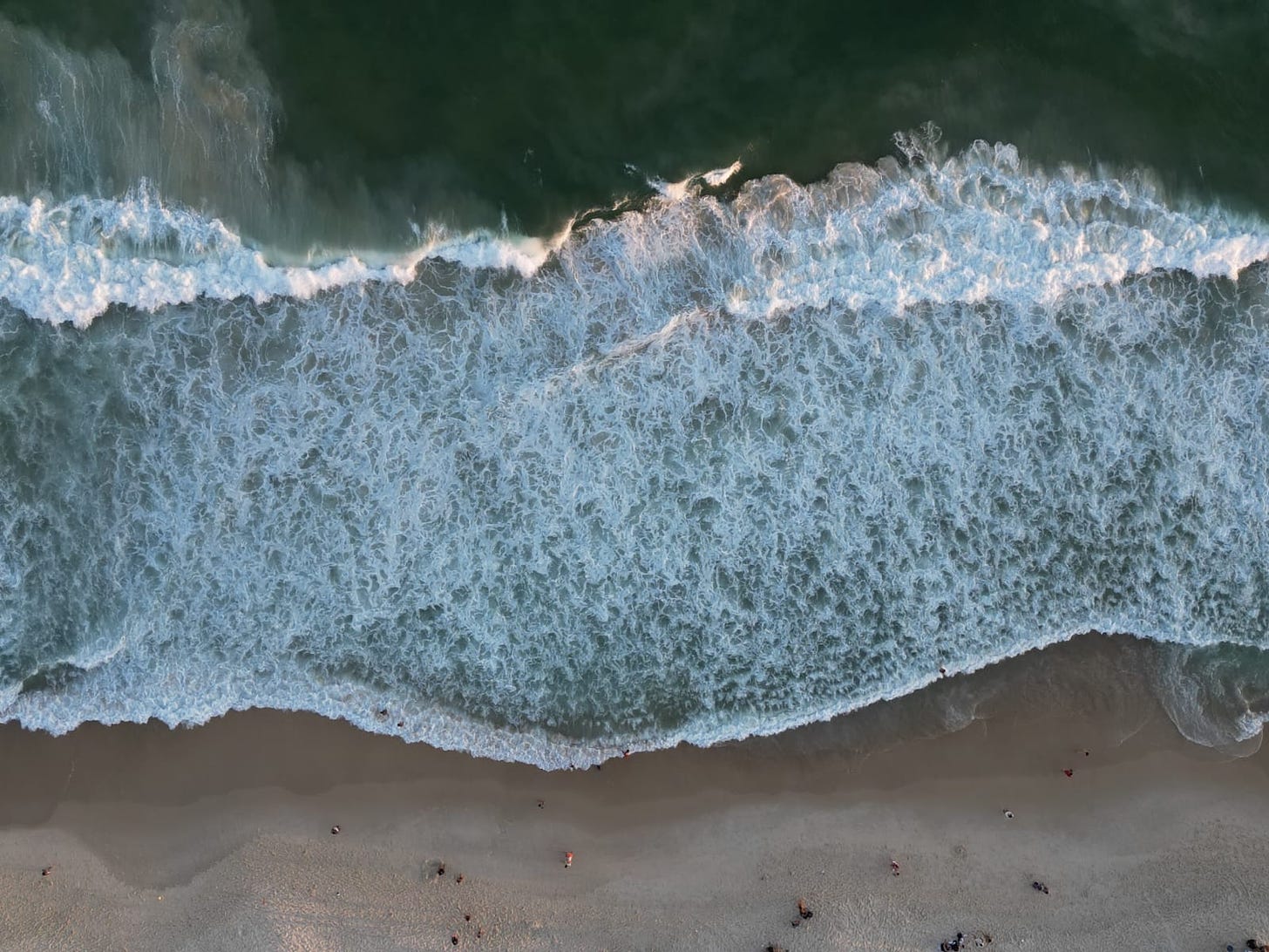 Fotografia de uma praia vista de cima - o mar, a espuma das ondas, a areia e pessoas caminando