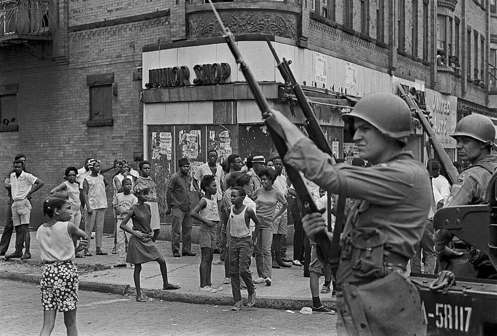 Eerie black and white photo, two National Guardsmen with bayoneted rifles in the air, across a corner store of about 30 black people, including children.