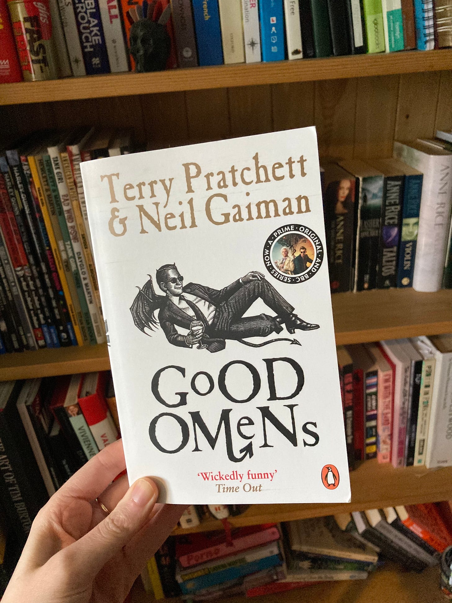 Good Omens by Terry Prachett and Neil Gaiman held in my hand, in front of my bookshelf