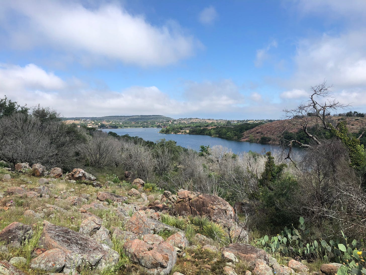 A photo from a trail at Inks Lake State Park depicting a blue body of water, blue sky, and a rocky embankment