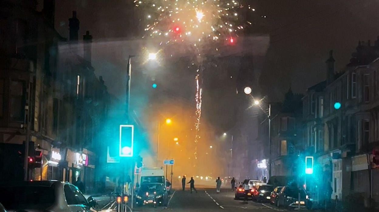 Fireworks set off from middle of road at night with figure standing in middle of road and explosions in sky between tenement windows