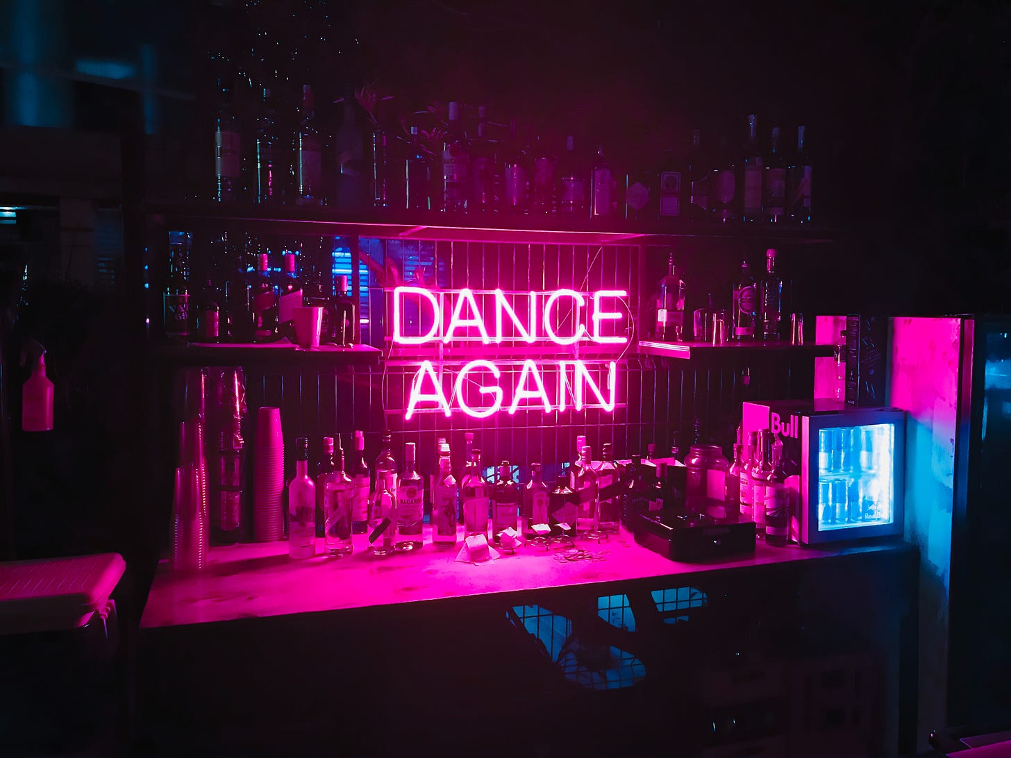 A sign in pink neon reads "Dance Again" set against a bar wall.