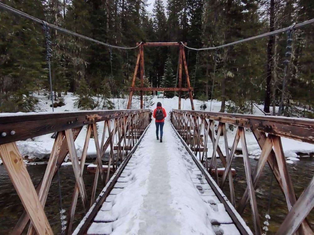 a woman in a red coat crossing a snow-covered wooden footbridge over a river 