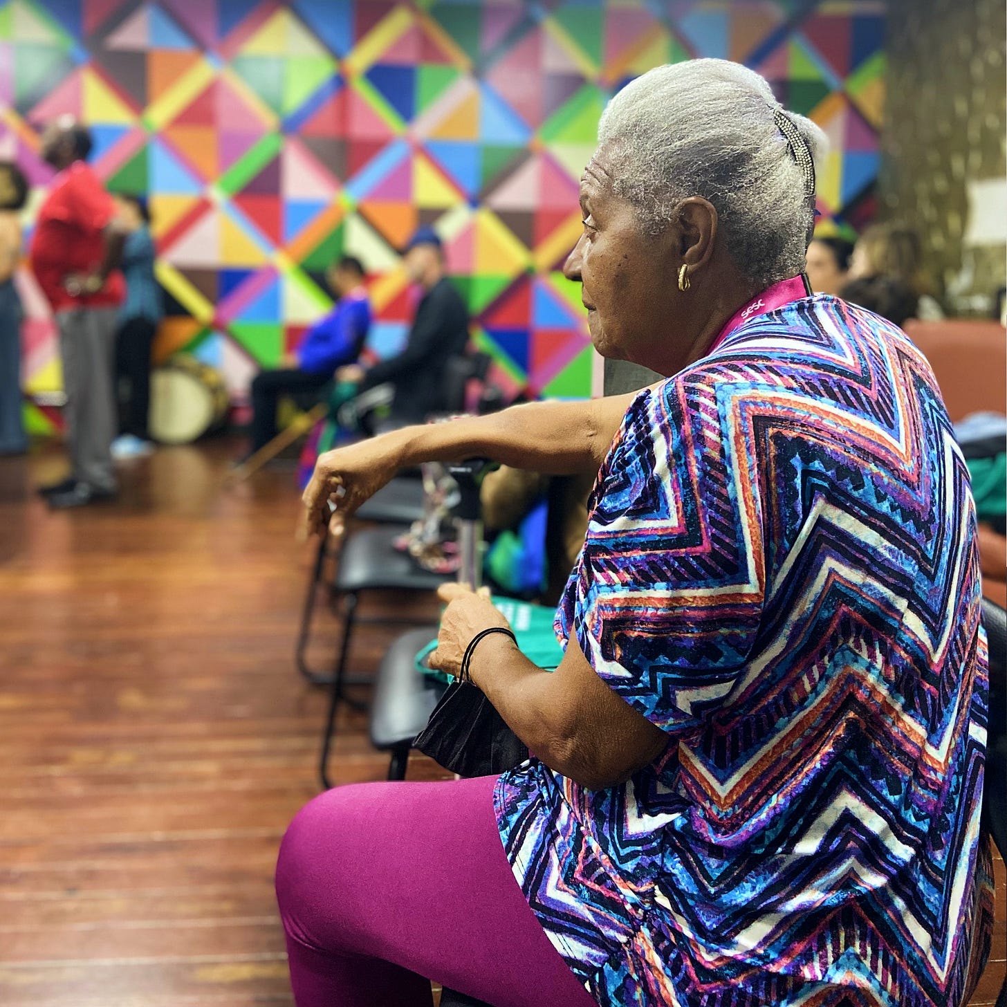 A foto mostra uma mulher mais velha, negra, de cabelos brancos, usando uma blusa colorida com estampa chevron e calças roxas, sentada e olhando para a esquerda do quadro. Ela está sentada em uma cadeira e apoia uma das mãos em sua bengala. Atrás dela e ligeiramente fora de foco, estão outras pessoas em uma sala com um padrão geométrico colorido nas paredes. É uma das atrizes do elenco de “A Velocidade da Luz”