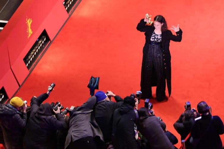 A woman laughs and makes a street gesture with her raised hand, holding up a gold statue in the other. She is on a red carpet facing a bank of photographers, and is wearing a black outfit.