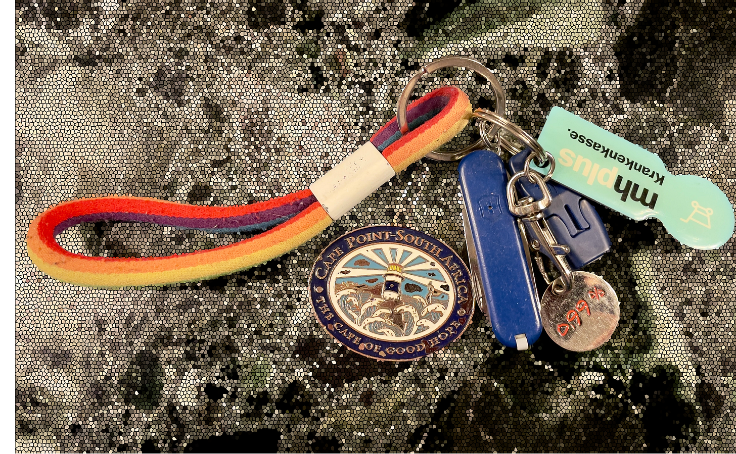 A minimal key set on a mosaic-textured surface, including a rainbow fabric loop, a Cape Point-South Africa medallion, a Swiss Army knife, a blue plastic key, and a German health insurance card keychain.