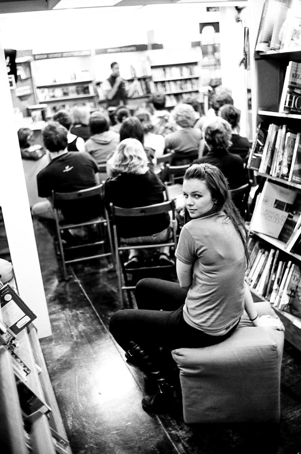 A poetry reading at a bookstore. In the foreground, Amber sits on a stool in the back of the audience. She turns her head to look at the camera behind her. In the background, slightly out of focus, is the audience watching Derrick perform. 