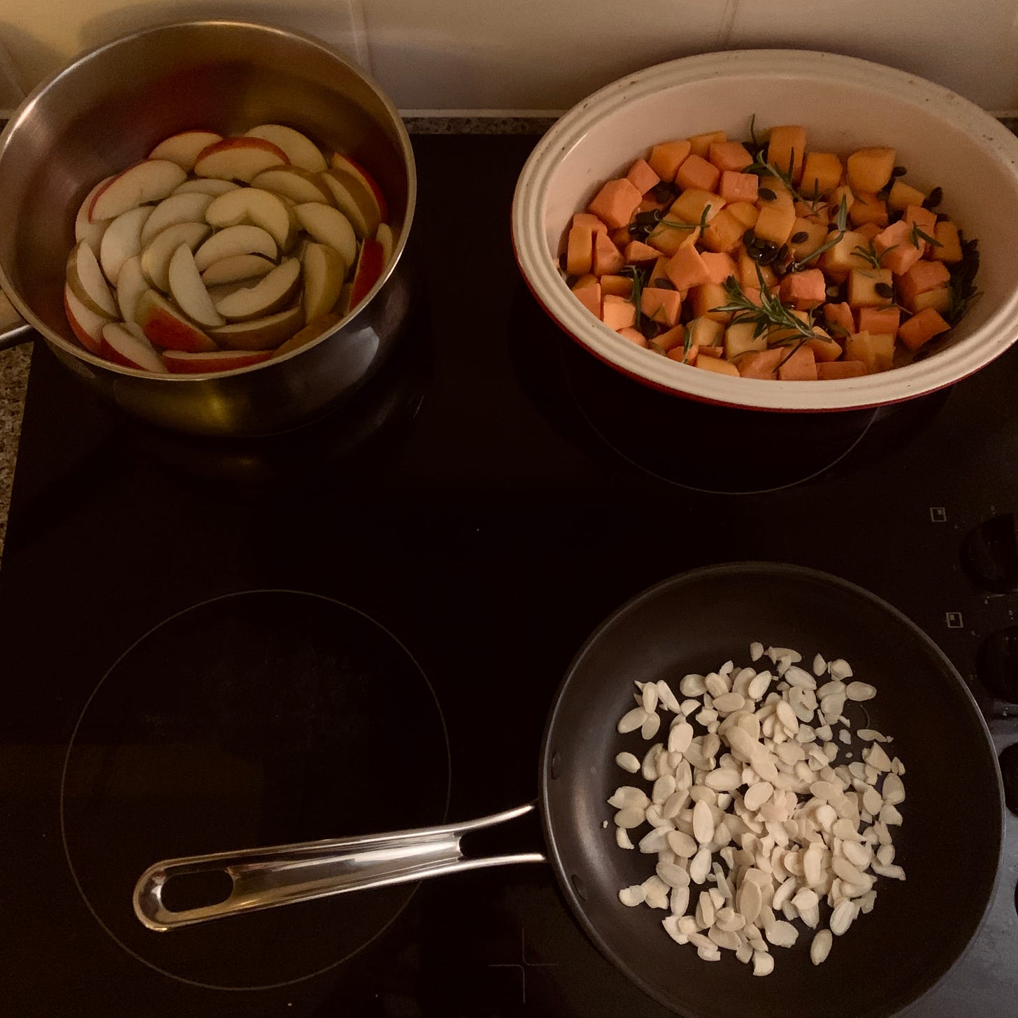 three pans on a hob, on is filled with apple slices, one with cubes of root vegetables and one with flaked almonds.