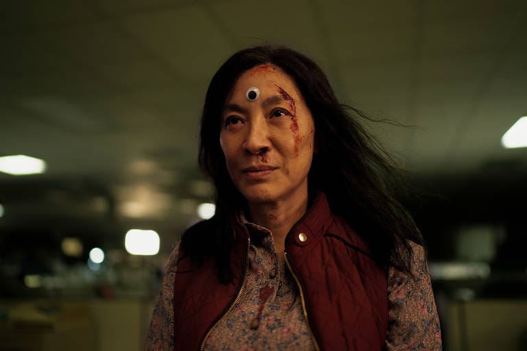 Michelle Yeoh, mulher malaio-chinesa de 60 anos, na personagem Evelyn - um colete vinho, uma camisa rosa, rosto tranquilo manchado de sangue, e um olho de brinquedo colocado na testa
