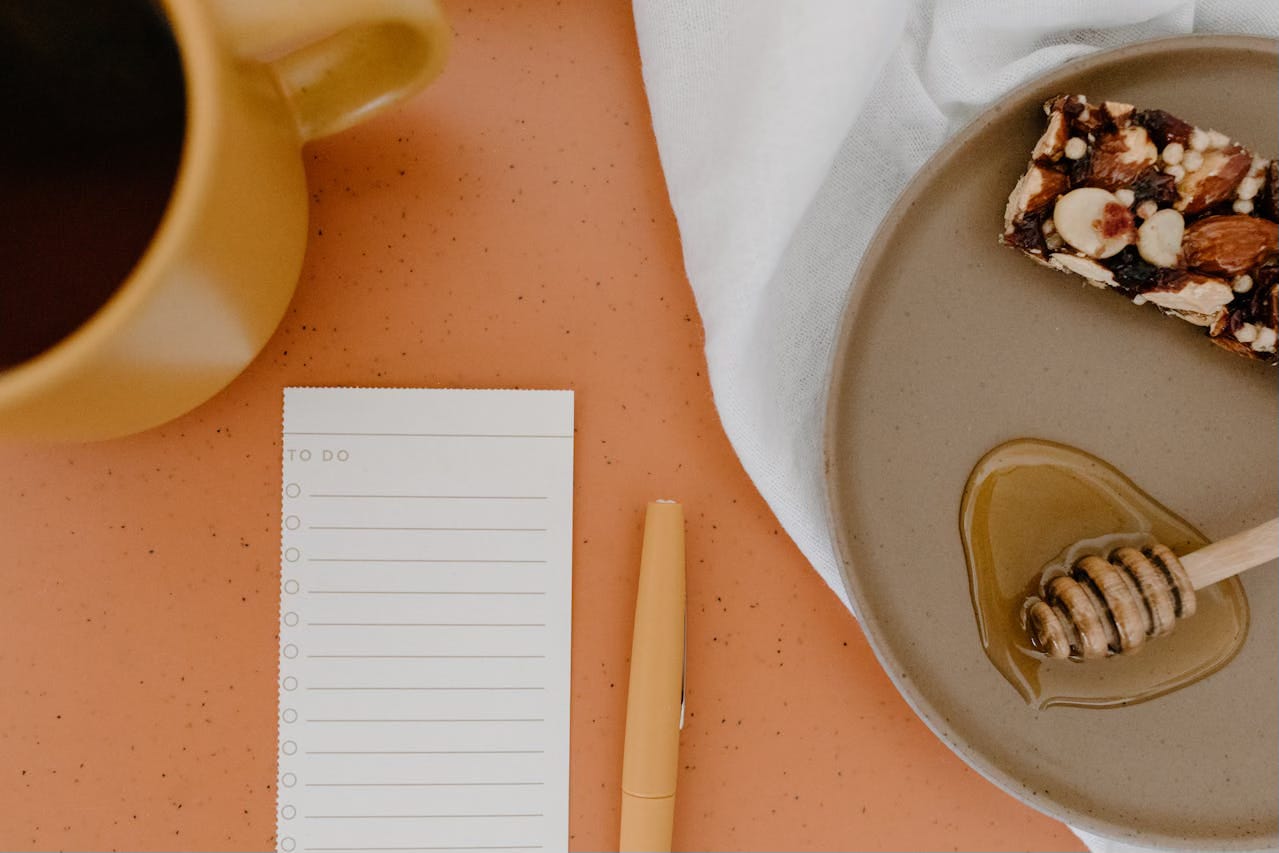 A to-do list pad sits on an organe tabletop next to a cup of coffee and a plate with a breakfast bar and honey at the end of a honey stick.