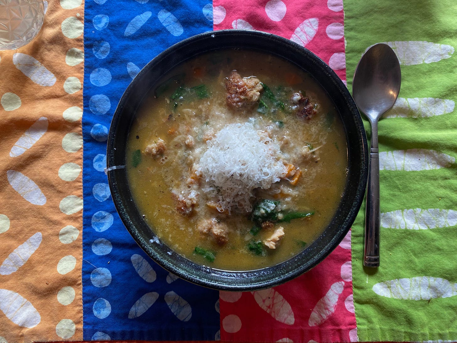 A ceramic bowl of Italian wedding soup, topped with a pile of grated Pamesan, on a colorful placemat.