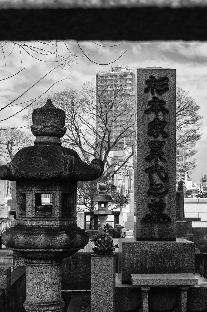 A monochrome winter photo of Yanaka cemetery in Yanaka, Tokyo, the burial place of Japan's last shogun, Tokugawa Yoshinobu. In the foreground are two large monuments, one in the shape of a helmeted man's head and torso on a sturdy pedestal, the other a large, triangular polished stone slab mounted on a pedestal with a small bench in front of it, and engraved in kanji. In the mid background are several bare trees, and in the far background is a large office block. The sky is cloudy and gray.