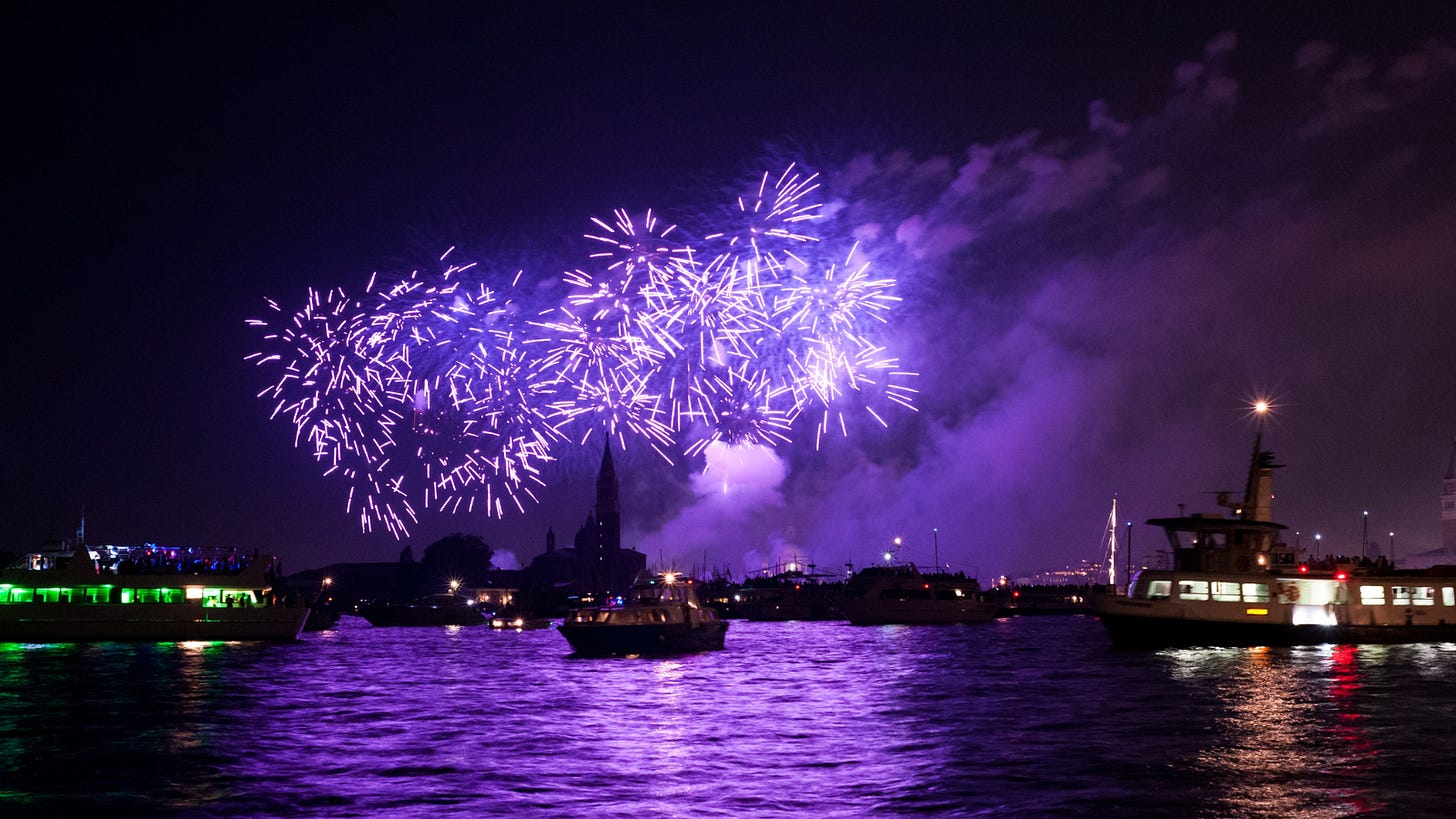 Purple fireworks in the night sky reflected in dark water where boats float