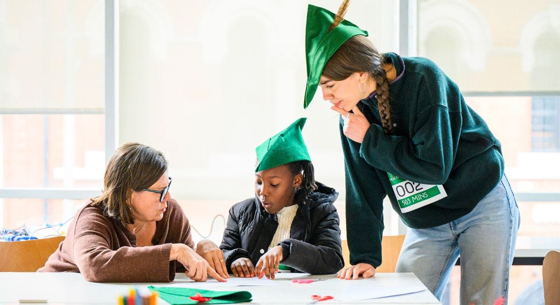 Three people sat at a white desk crafting green felt Robin Hood hats, with windows behind them