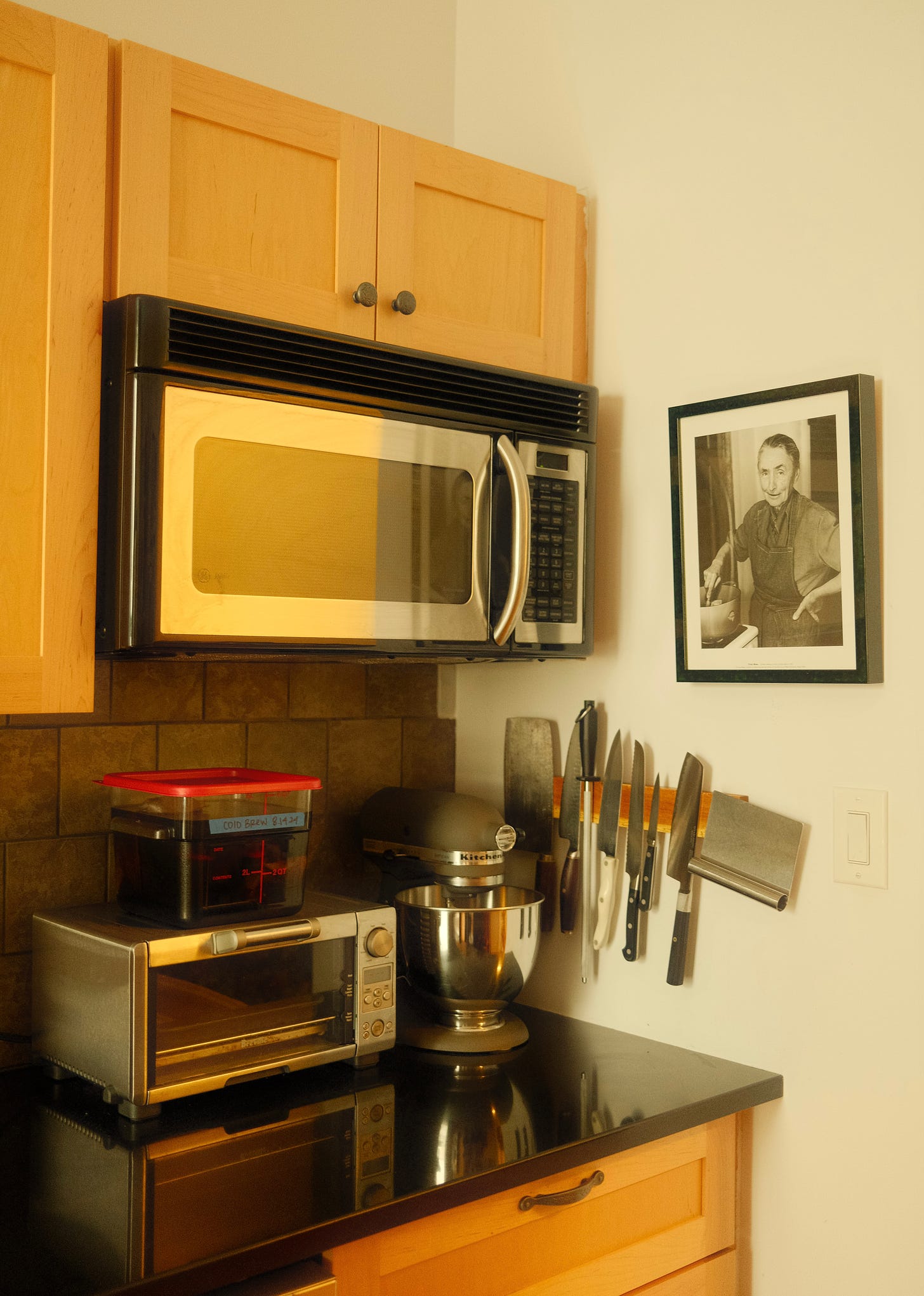 Kitchen gadgets on a counter top. Microwave fixed to the wall under wooden cupboards.