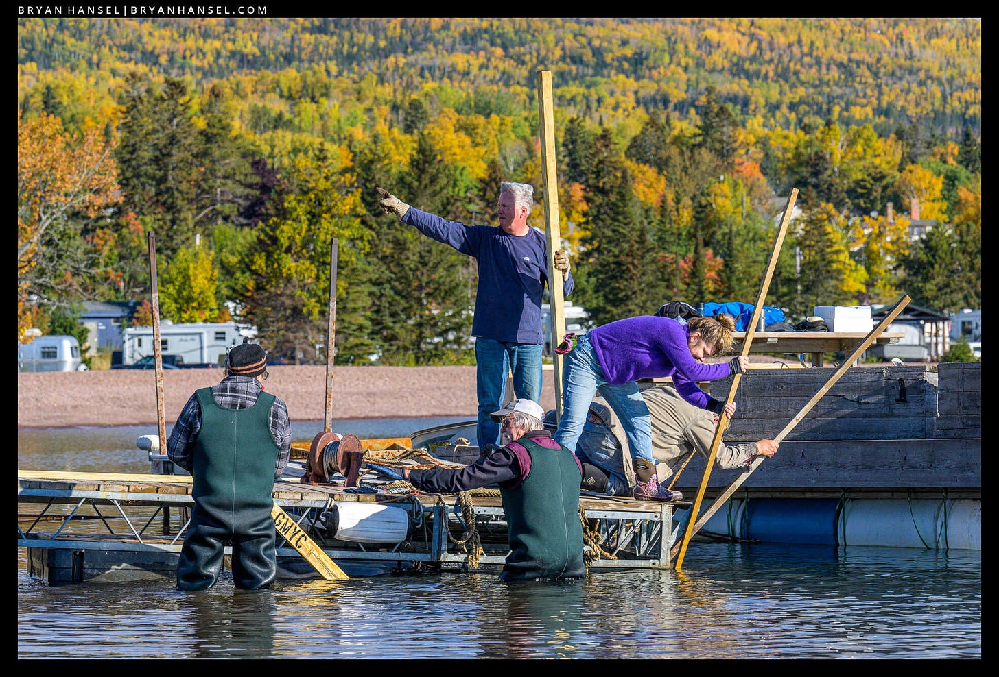 Two people pushing a dock in the water. Three people stading on the dock and two pushing something underwater with sticks. There's a third man pointing towards shore. Fall colors in the background.