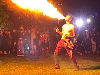 A man, naked from the waist up, breathes out a giant wave of fire as he performs firebreathing in a park at night, a crowd of adults and children standing watching at a safe distance.