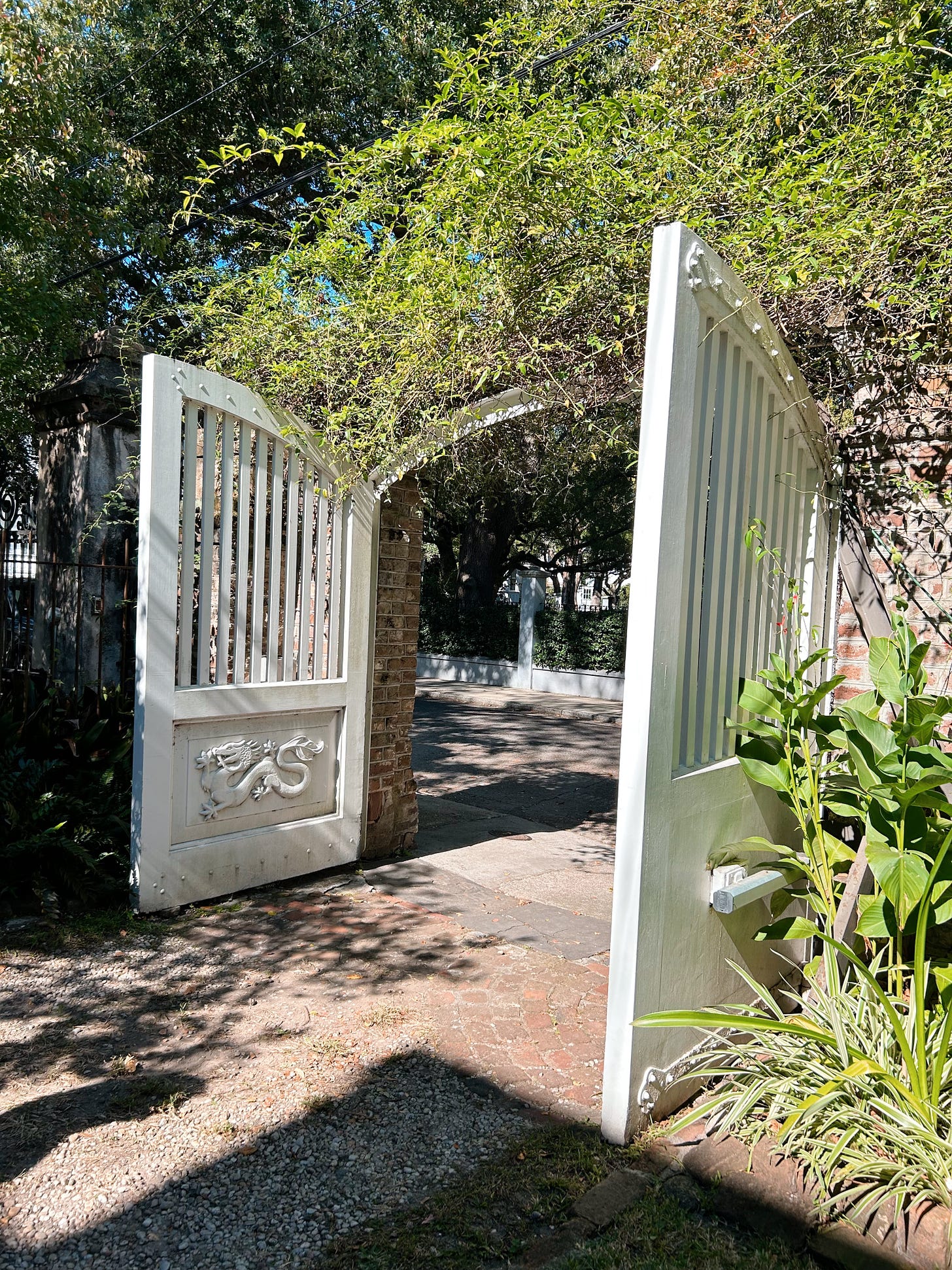 White gates carved with the dragon emblem from the Wheel of Time. Vines and green plants surround and shade the driveway.