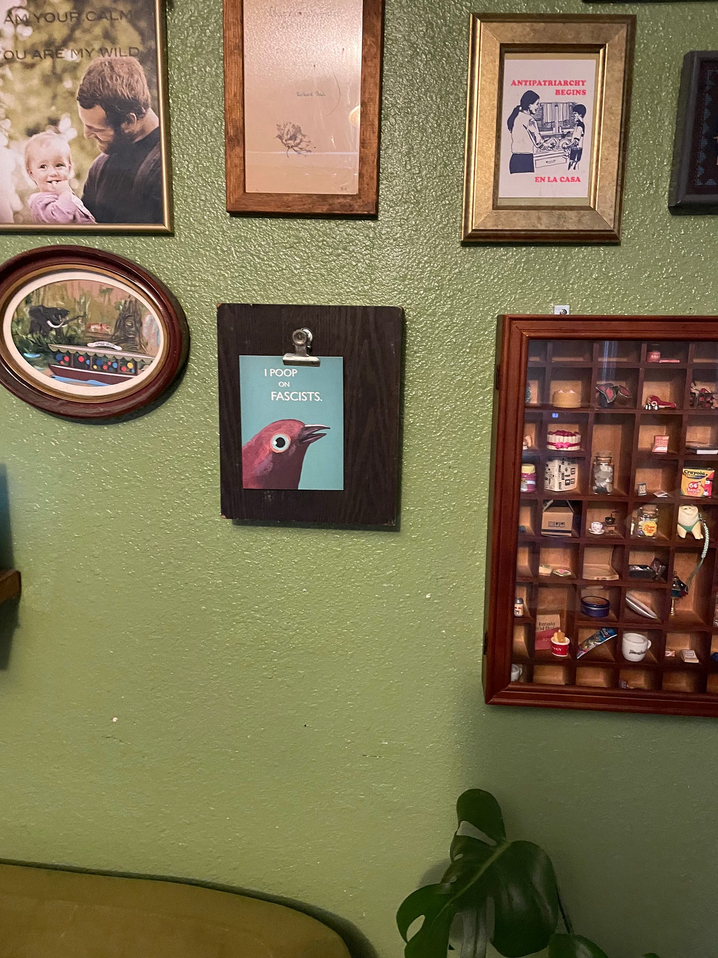 a green wall with a bunch of various pictures hanging on it, mostly with wooden or gold frames. There is also a wooden miniatures cabinet. One a wooden plaque there is a small blue card with the paintnig of a bird on it. In small white text it says "I poop on fascists"