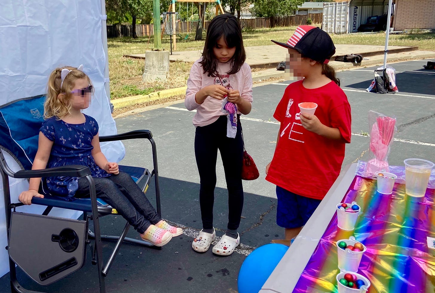 two children standing and one girl sitting at an outdoor booth