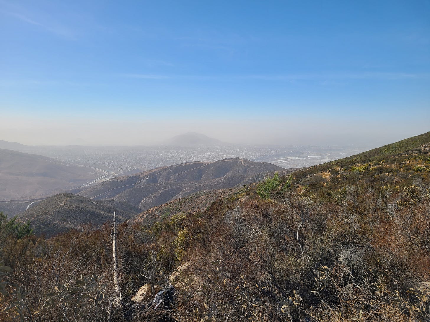 a brush-filled mountain landscape with the border wall and Tijuana in the distance down the mountain