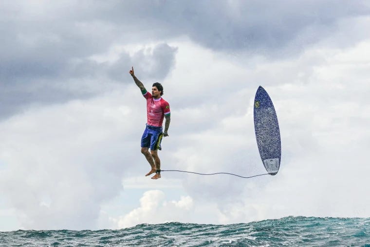 A male-presenting surfer appears to be floating many feet above the ocean's surface holding up his index finger. His surfboard is also floating in the air parallel to him.