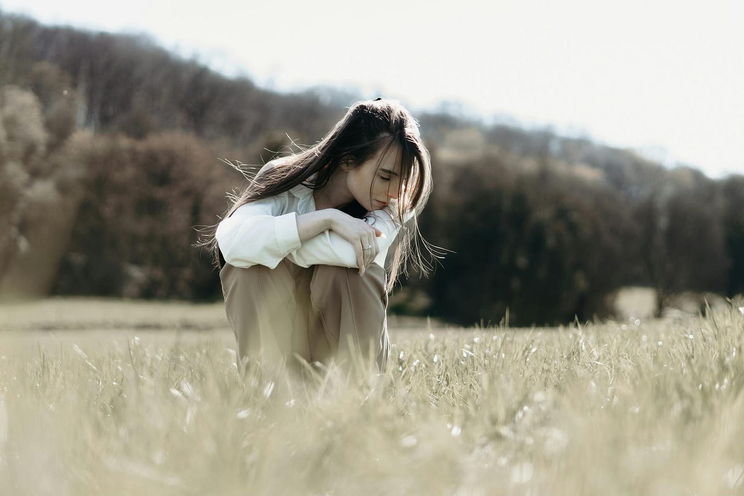 A woman with long hair and dressed in being sits in a dry field looking to the side and downward.