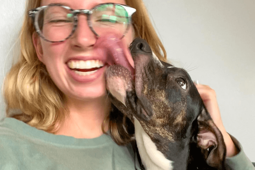 Haley, a young blonde woman, takes a selfie with her foster puppy Mystic, a black and white mixed breed, licking her face