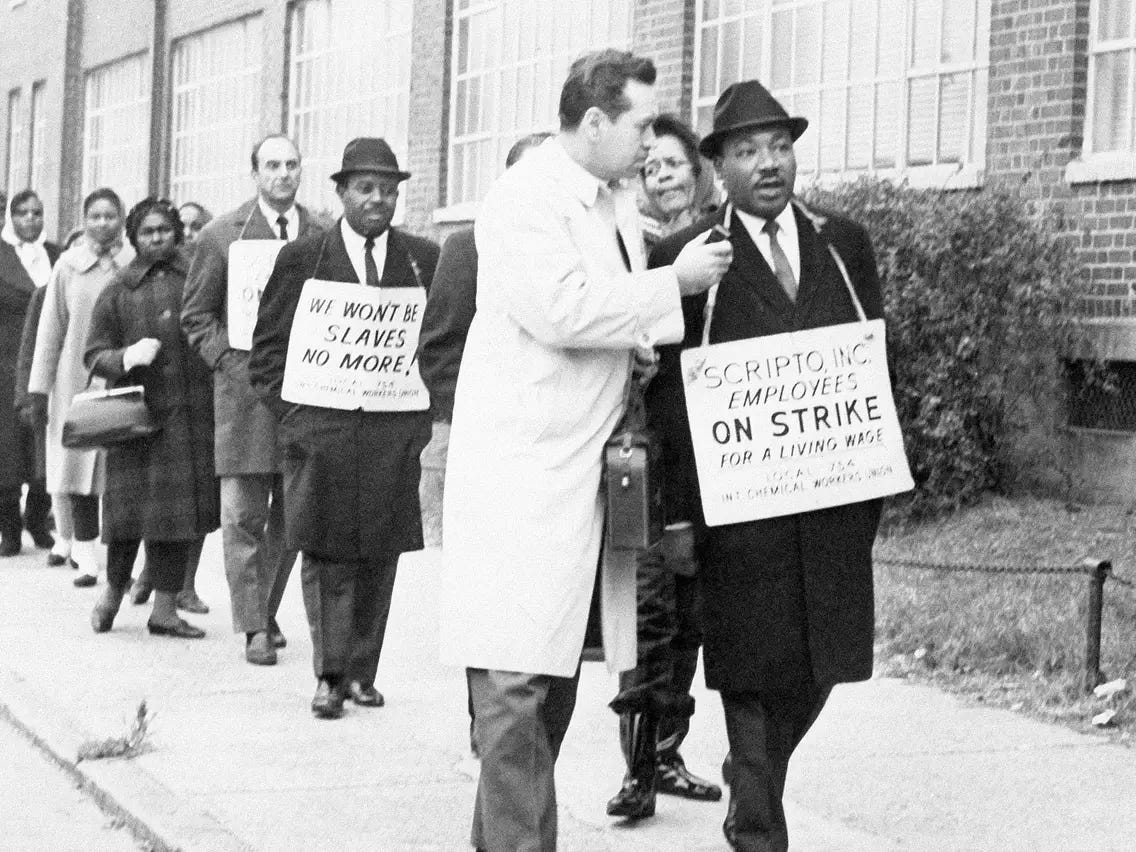 Dr. Martin Luther King on a picket line during the 1964-65 Scripto strike in Atlanta, Georgia. He is at the front of the picket line, wearing a sign, and being interviewed by a reporter.