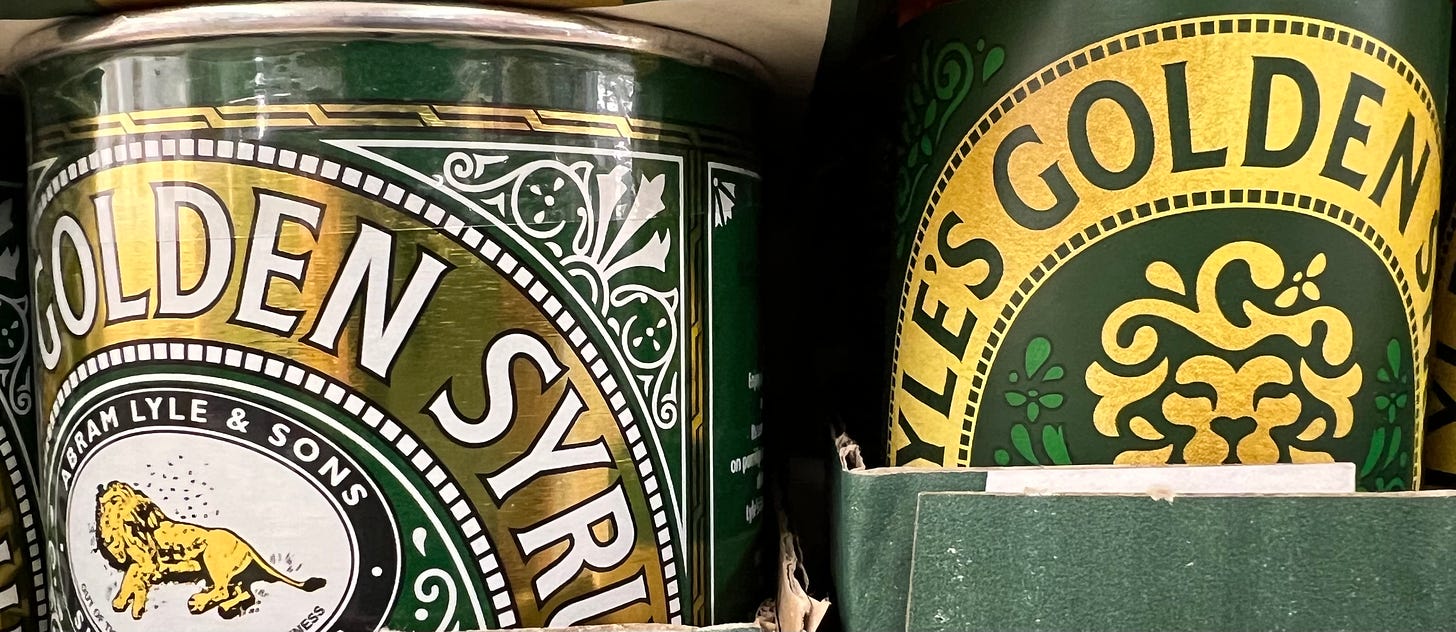 Two containers of Lyle's Golden Syrup on a shelf. A metal tin with a Victorian brand design featuring a dead lion, and a plastic bottle with a modern design featuring a (presumably) living lion.