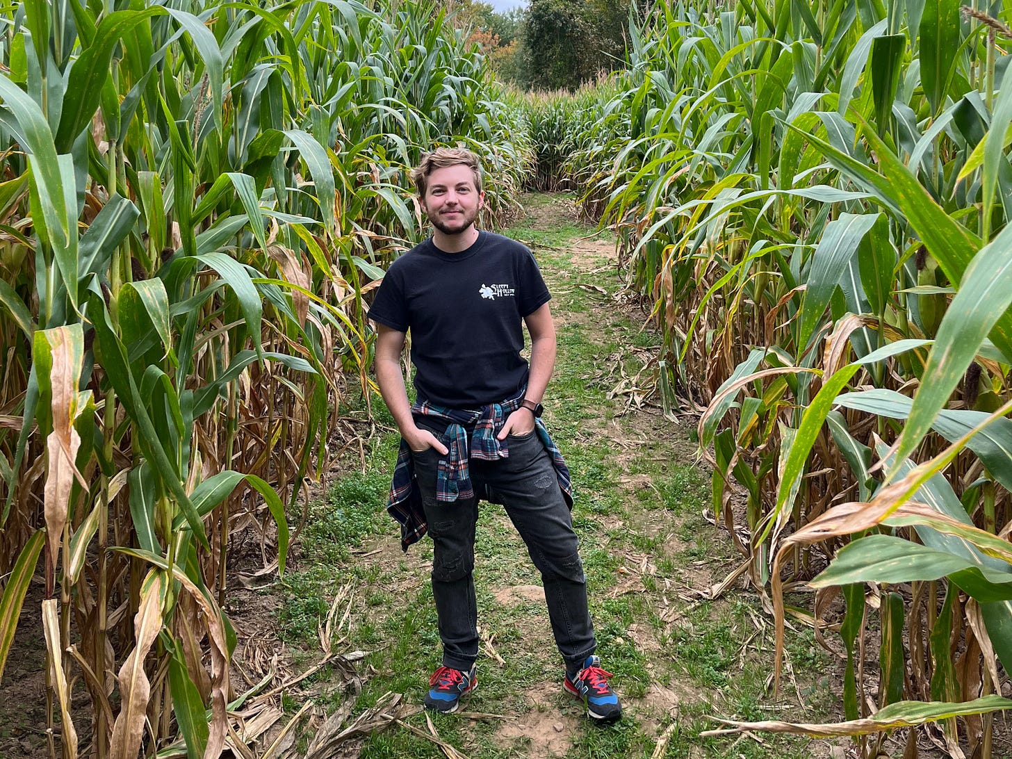 Jack stands in the middle of a row of corn with his hands in his pockets, looking happily at the camera