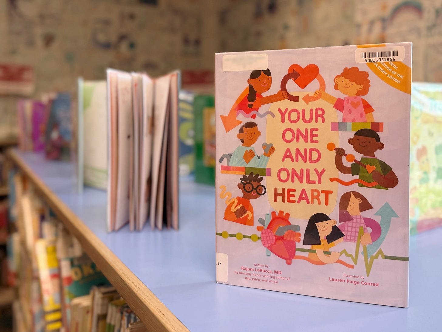 The picture is from the top of a row of shelves in the children's library in Huntington Beach, CA. Several books are on display but the only visible cover is Your One and Only Heart, a colorful picture book about the human heart.