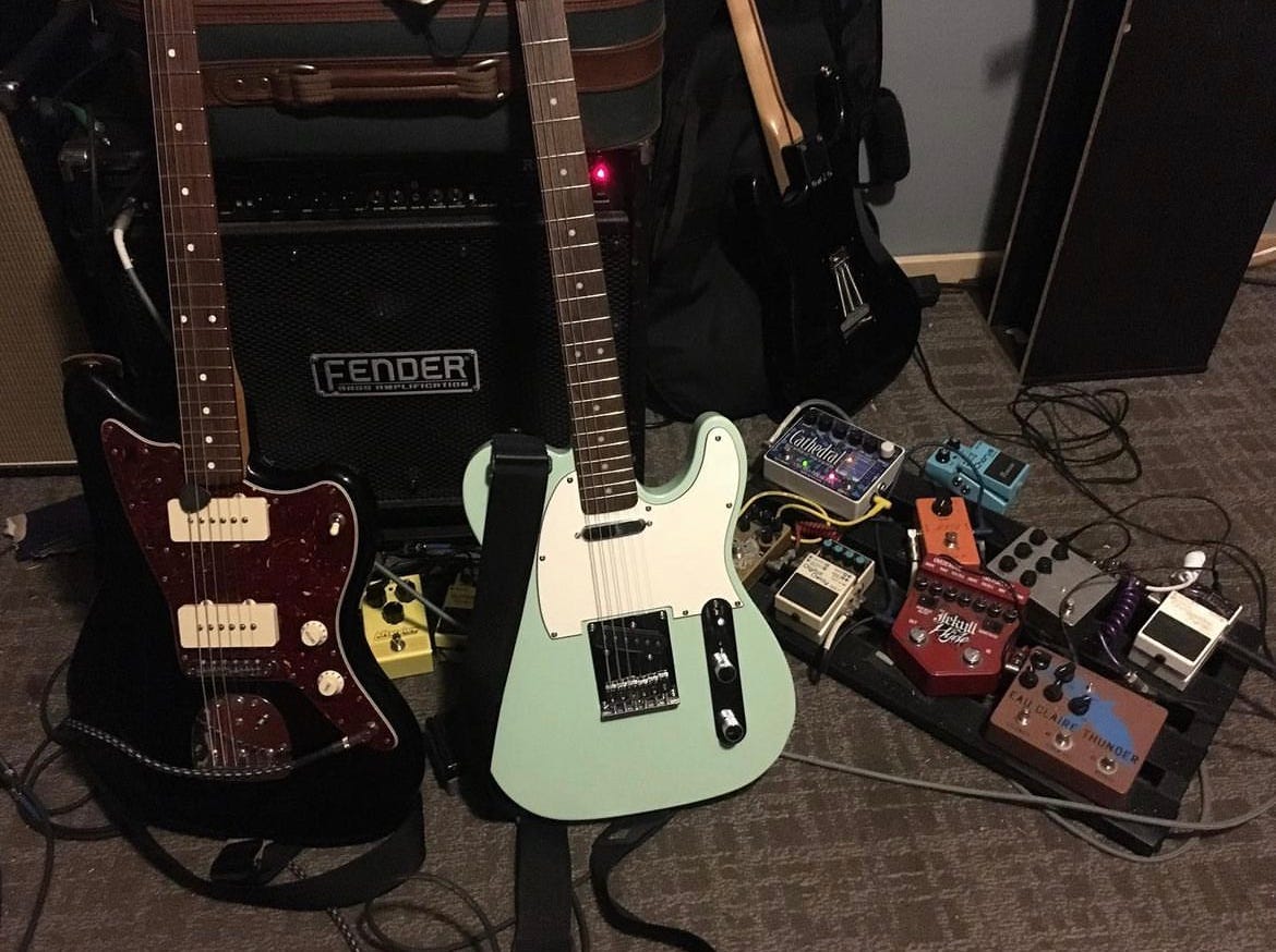 a black Fender Jazzmaster and blue Squier Telecaster rest against a 12" Fender combo amp. Next to the guitars is a pedalboard, but it's someone else's pedalboard, and I don't know what kind of pedals they are. The carpet is surprisingly clean for a practice space.
