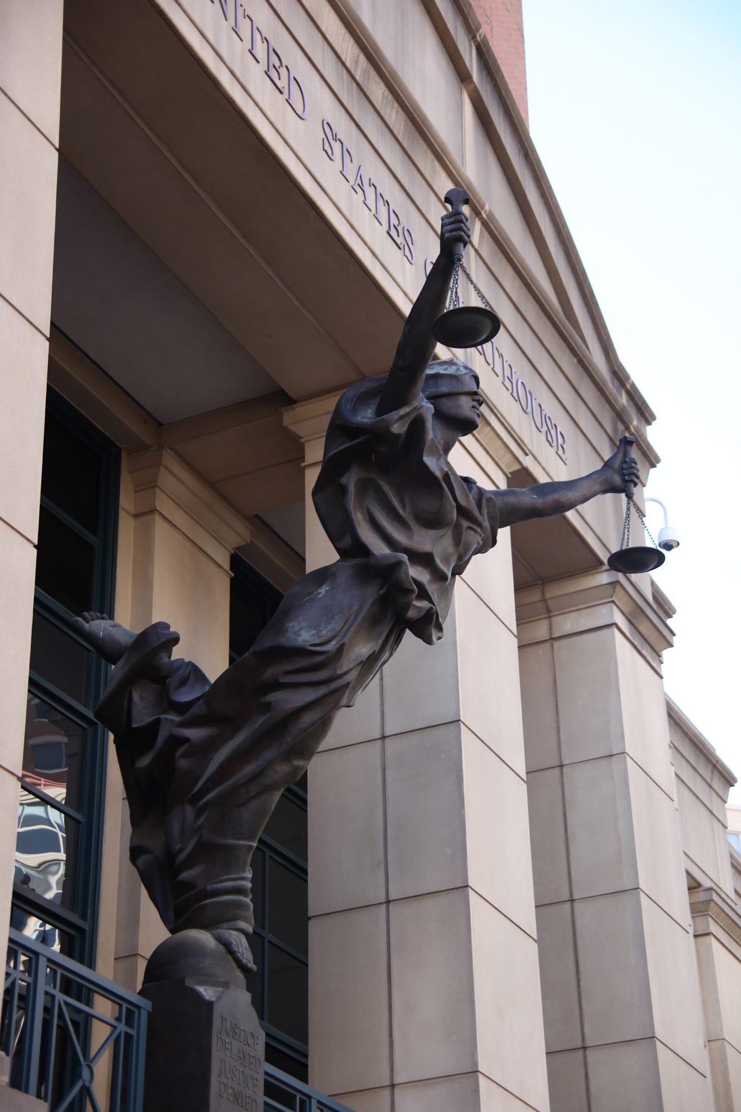 Dark-colored statue of a blindfolded woman balancing on one foot, holding scales in each hand. She is standing on a pedestal that has the wording "Justice delayed justice denied" on it. She is in front of a courthouse. The wording "UNITED STATES COURTHOUSE" is visible, but partially obscured by her, behind her. She is leaning forward so that her balance appears precarious. Dark-colored statue of a blindfolded woman balancing on one foot, holding scales in each hand. She is standing on a pedestal that has the wording "Justice delayed justice denied" on it. She is in front of a courthouse. The wording "UNITED STATES COURTHOUSE" is visible, but partially obscured by her, behind her. She is leaning forward so that her balance appears precarious.