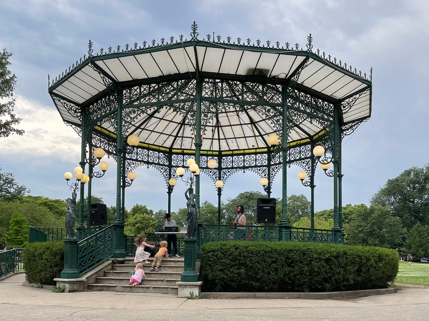 A green and white gazebo surrounded by hedges and trees. Under the gazebo are two musicians. There are three toddlers climbing the stairs of the gazebo.