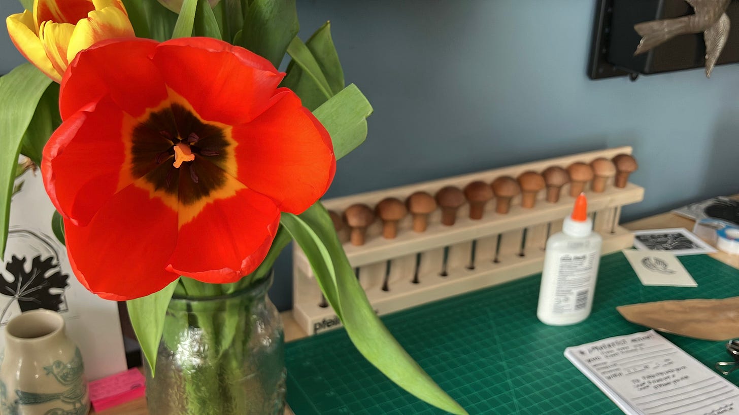 an orange-red tulip opens up to show off its dark interior. It's on my workbench, where there's a green cutting mat and printmaking supplies of various kinds.