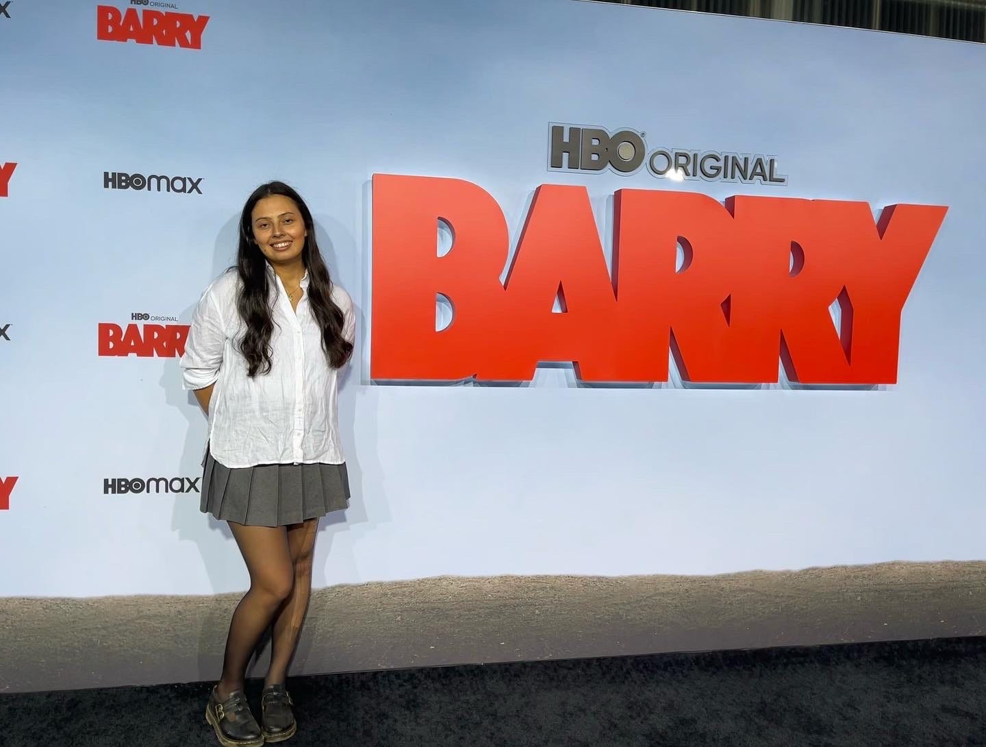 Nicole Saucerman, a woman with long, dark hair, a white shirt, and a gray skirt, standing next to a red sign that says "BARRY"