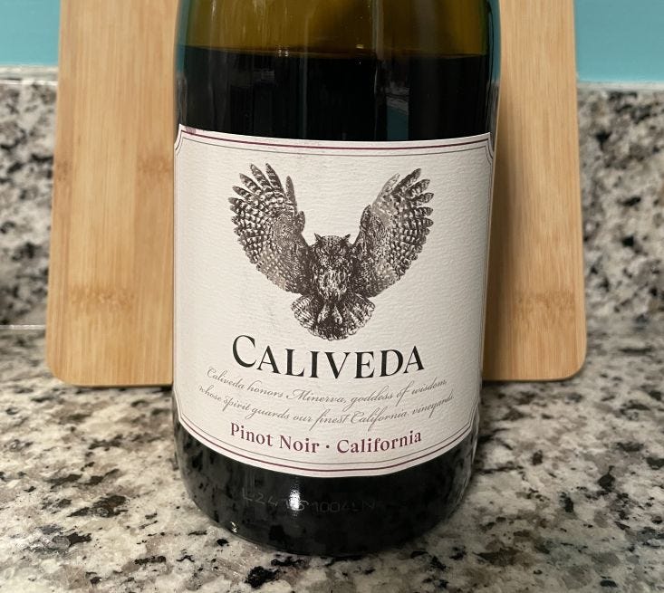 Bottle of red wine on granite counter in front of cutting board and blue wall; wine label has on owl on it.