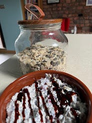 In the foreground, is a brown ceramic bowl of cereal. It's white, full of yoghurt instead of milk, and seeds and nuts poke out. On top, there's a brown, treacle-like drizzle. In the background, on the worktop behind the bowl, is a large glass jar with a wooden lid. Inside, there are nuts, seeds, and oats, all mixed together.