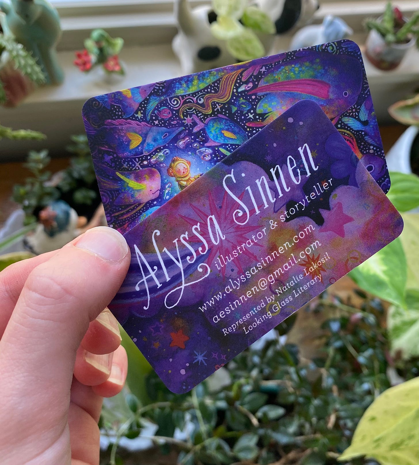 Hand holding two purple colored business cards with the artists' contact information, backdropped by many houseplants.