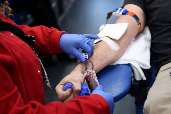 A close-up of a phlebotomist in a red jacket and blue gloves assisting a person donating blood from the right arm.