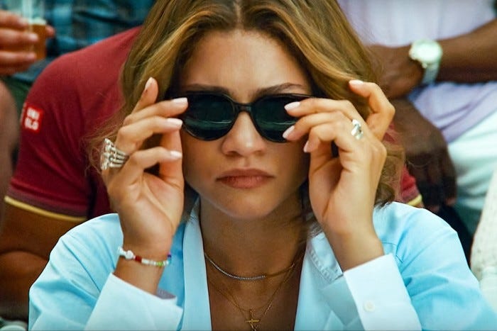 A close-up view of a young woman in dark glasses and pale blue open-neck top watching a tennis match