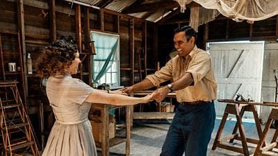 A woman and a man hold hands as they dance in a wooden warehouse. They grin as they spin around. 